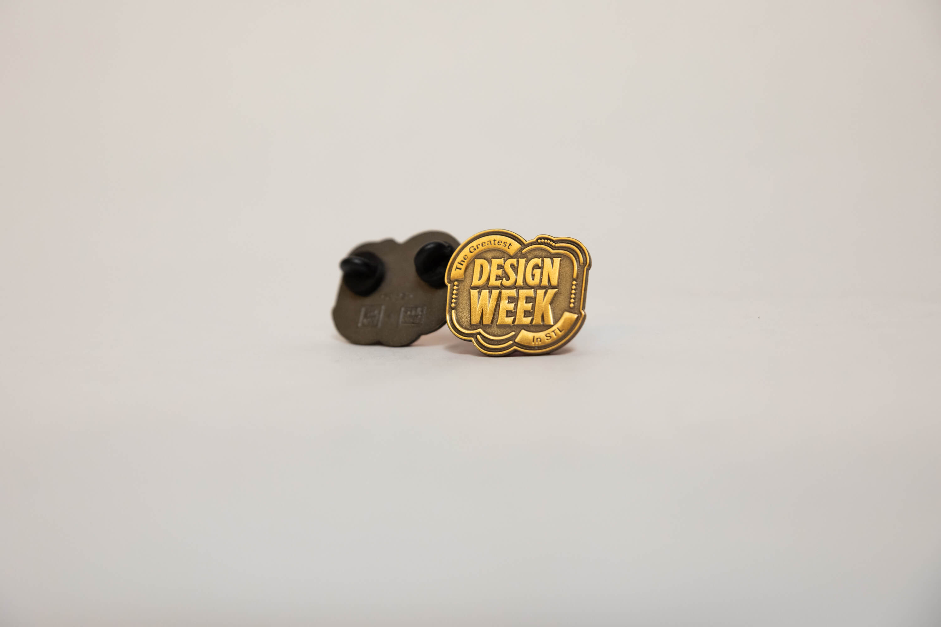 Enamel pin with gold lettering that reads 'The Greatest Design Week in STL' and a black pin backing.