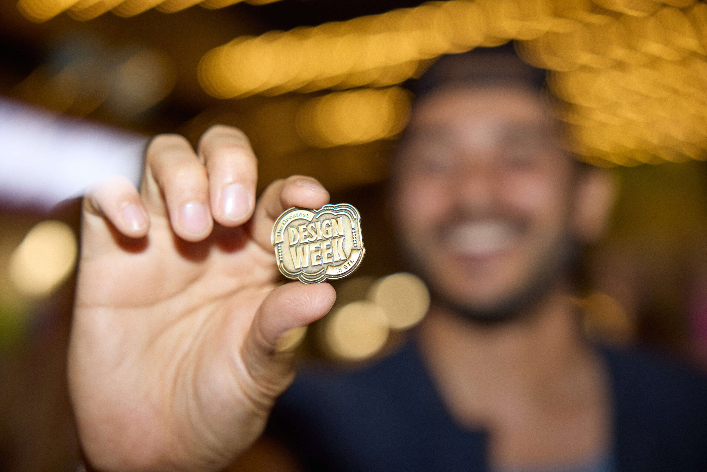 Person holding a pin that reads 'The Greatest DESIGN WEEK in STL' against a background of blurred warm lights.
