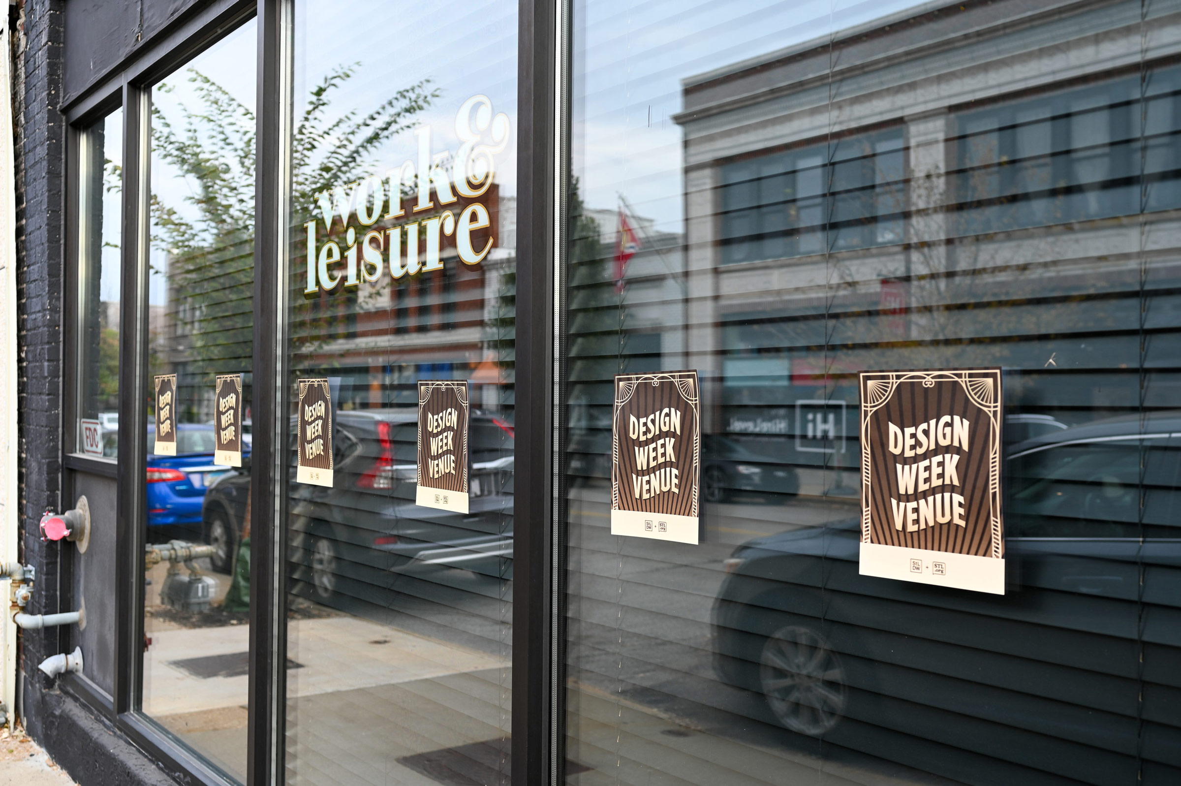 Glass window of a building with reflections of street and cars, showing multiple 'Design Week Venue' signs and the text 'work & leisure'.