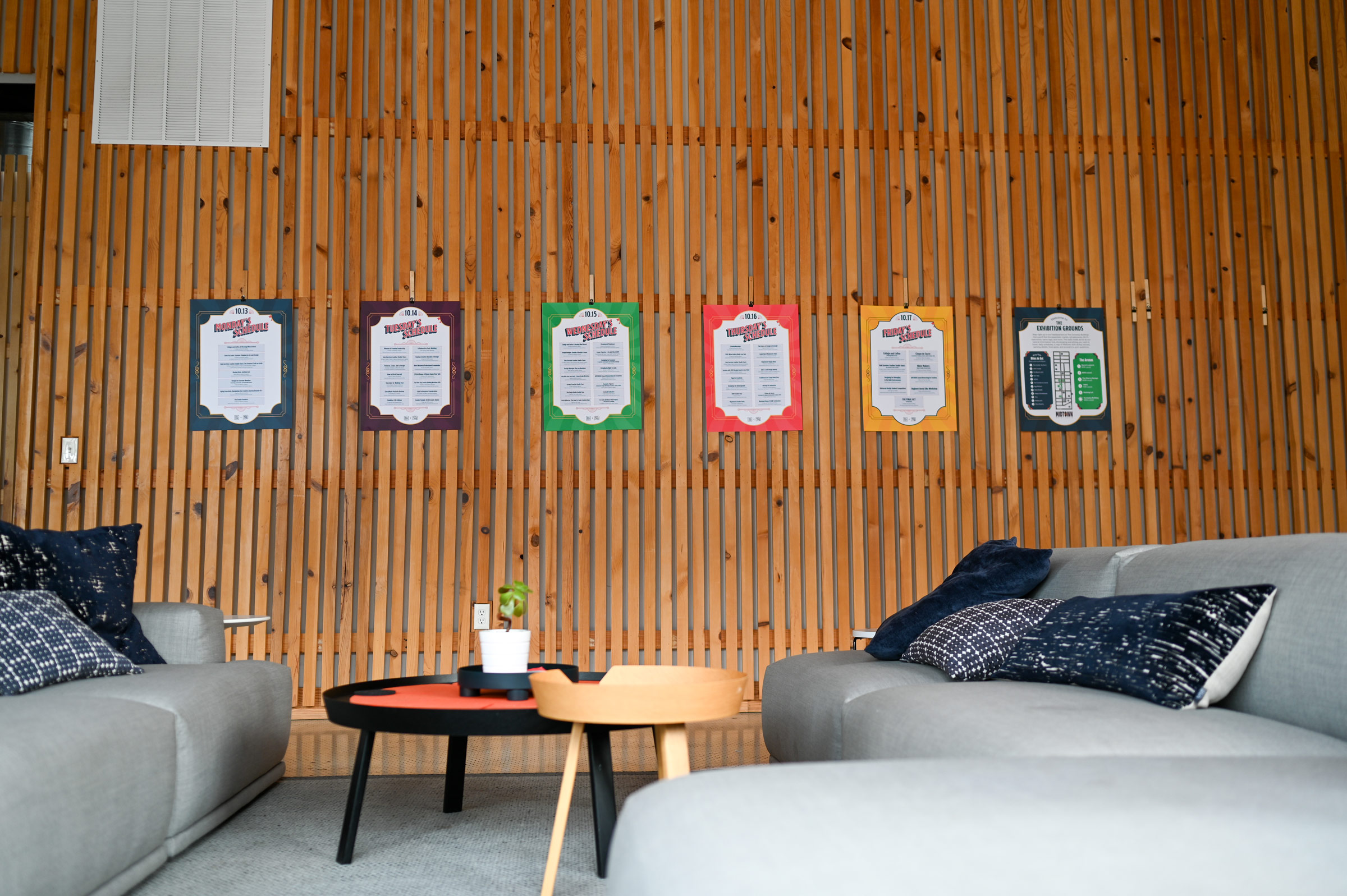 Modern lounge area with two gray sofas, black and patterned pillows, a round black coffee table with a small potted plant, and colorful schedule posters on a wooden slatted wall.