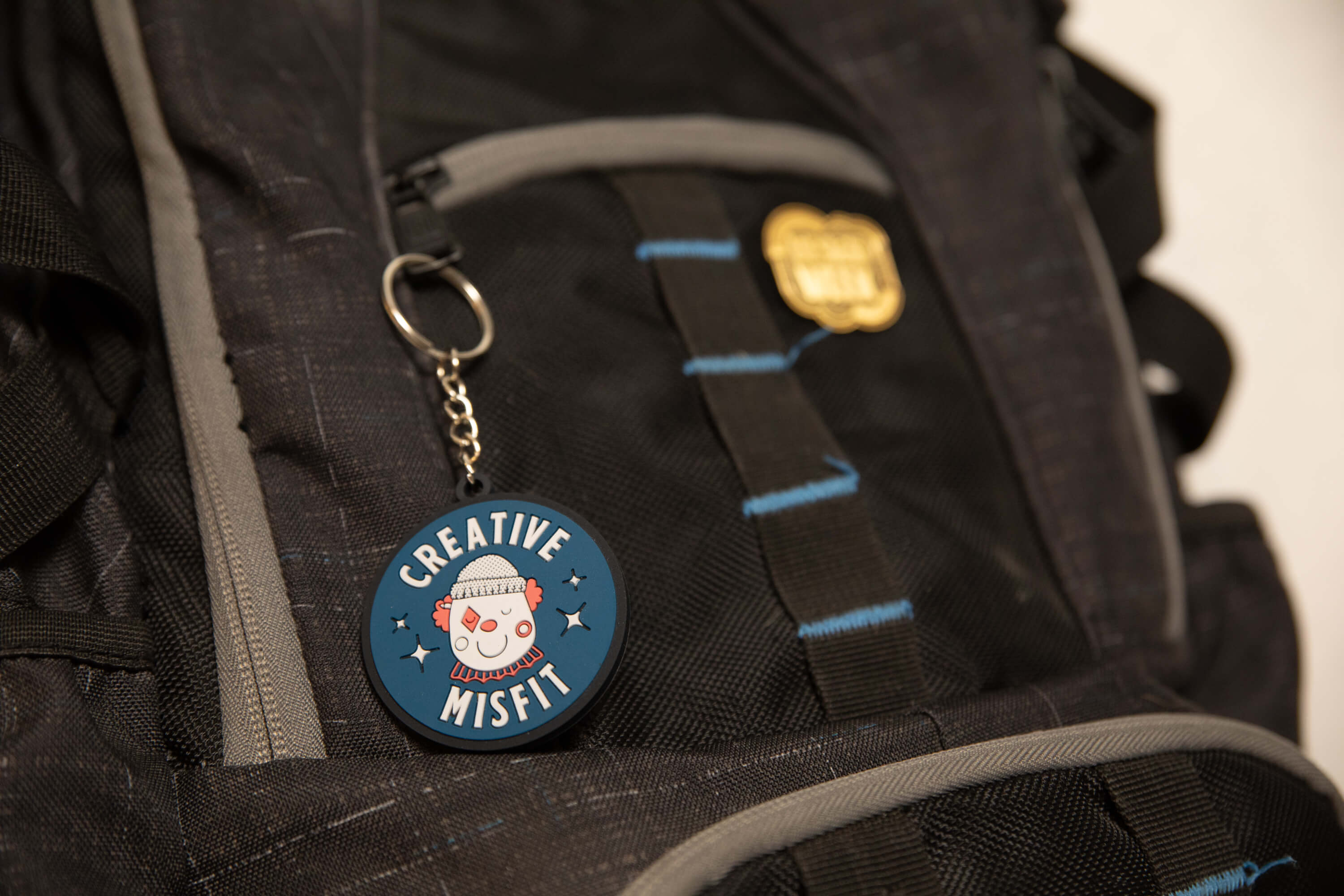 Blue circular keychain with a clown face and the words 'Creative Misfit' attached to a black and gray backpack.