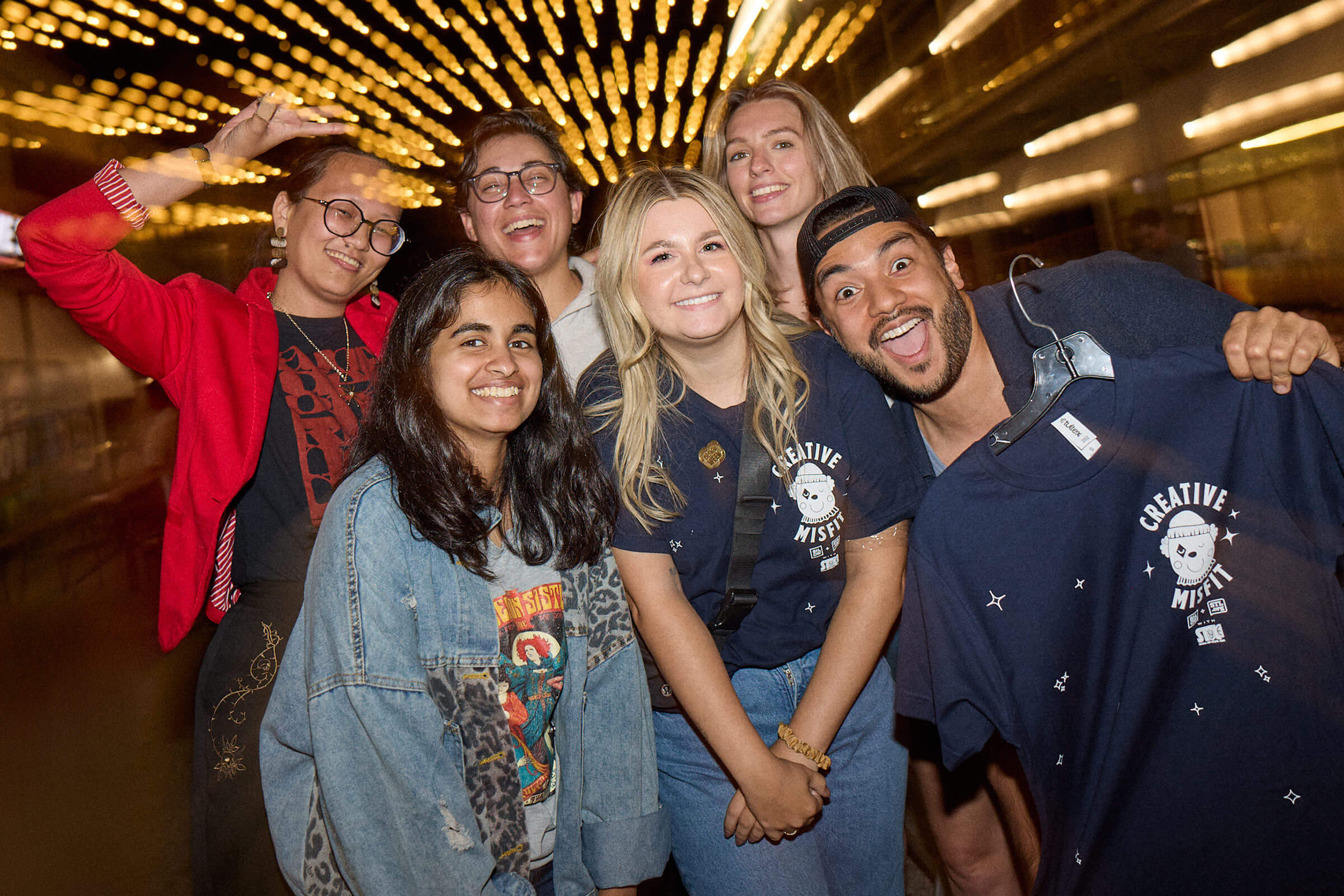 Group of six smiling young adults posing under warm string lights, with one man holding a 'Creative Misfit' t-shirt.