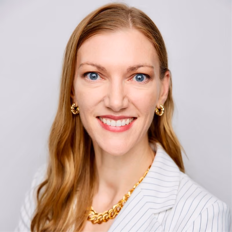 Smiling woman with long blonde hair wearing gold jewelry and white pinstripe blazer.