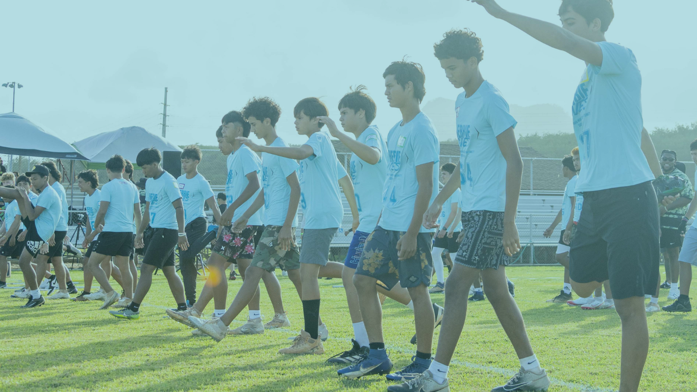 Group of young athletes wearing light blue shirts reading “Herbie Gives,” walking in formation across a grassy field during an outdoor sports camp, with bleachers and tents in the background.