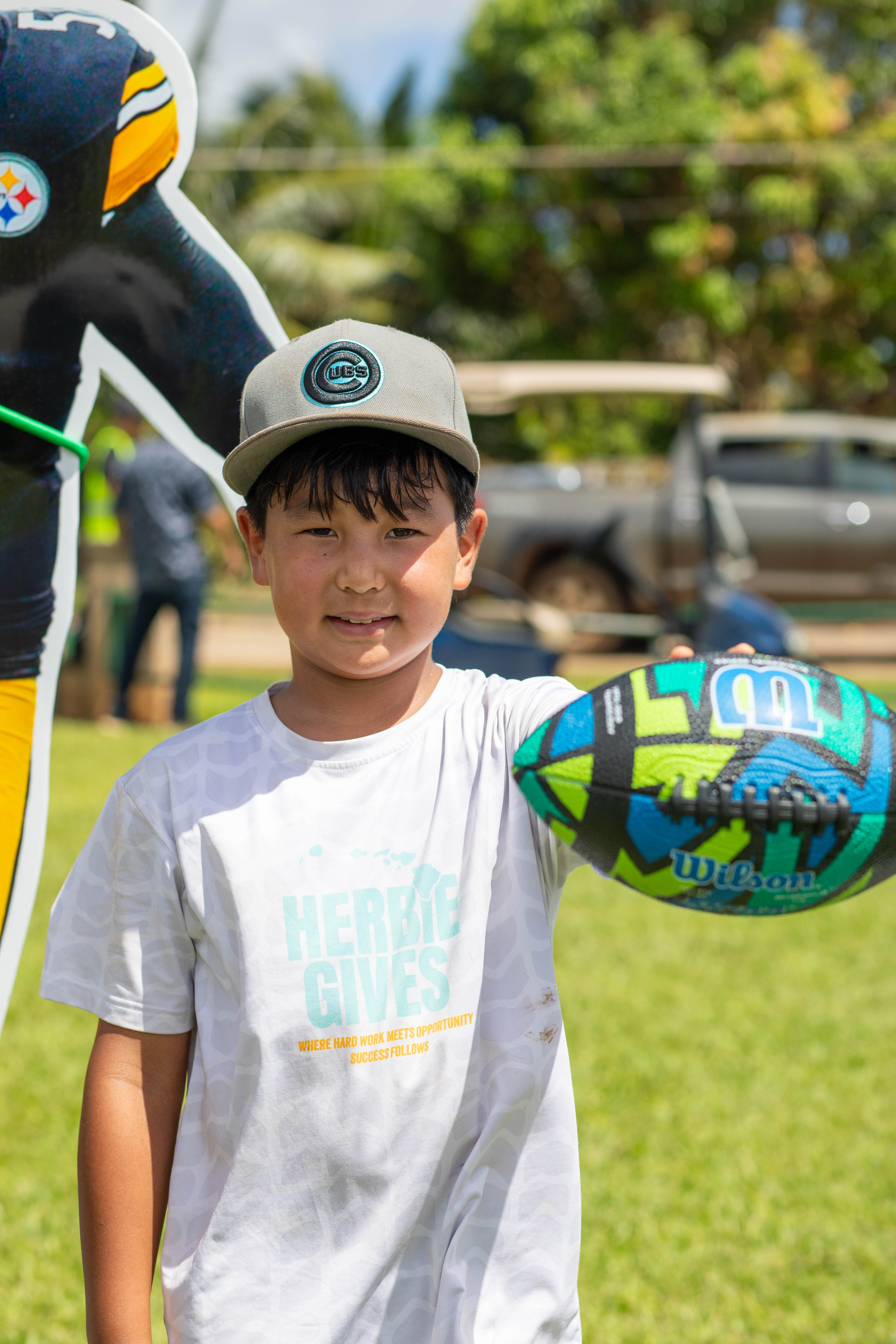 A boy in a HerbieGives shirt holds out a football and smiles