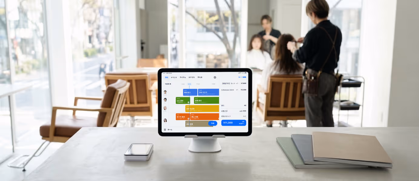 美容室での予約システムと連携したPOSレジアプリが表示されたタブレット端末が置かれたカウンター。背景には施術中の美容師と顧客。