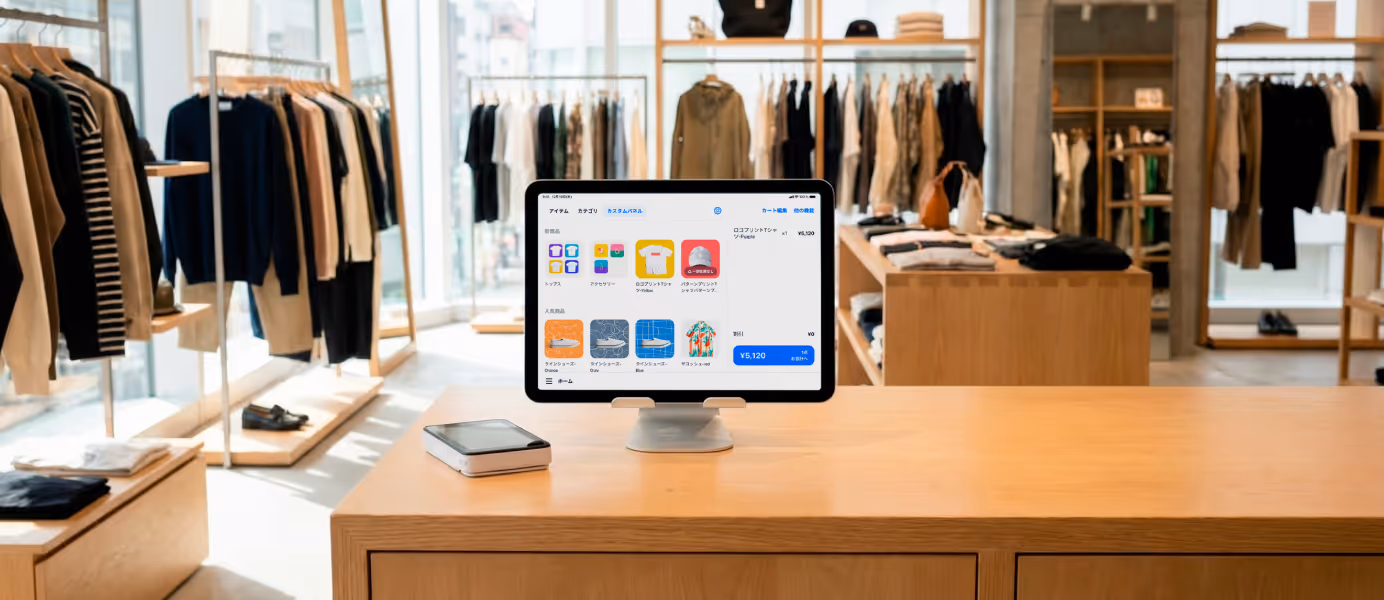 洋服店(アパレル店)のカウンターに置かれたPOSレジアプリの画面を表示したタブレット端末と決済端末。背景にはさまざまな衣類が並ぶ店内。