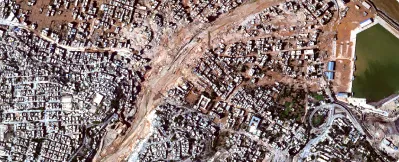 Aerial view of a densely built urban area with a large cleared diagonal path and a small water reservoir on the right.