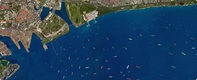 Satellite view of a coastal city with numerous ships anchored or sailing in the harbor and open sea.