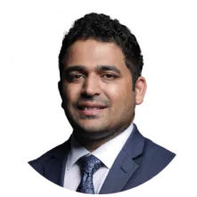 Headshot of a man with dark curly hair wearing a navy suit, white shirt, and patterned tie, smiling against a white background.