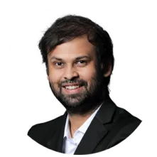 Smiling man with dark hair and beard wearing a black blazer and white shirt against a white background.
