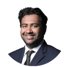 Smiling man with short curly hair wearing a dark suit, white shirt, and black tie against a white background.
