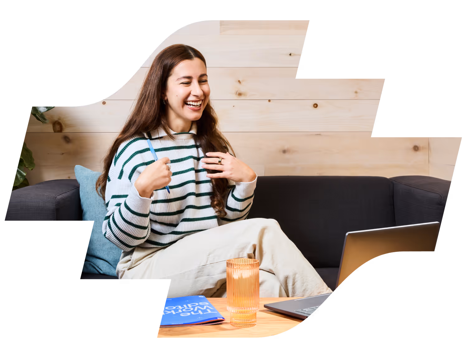 Smiling woman in striped sweater holding pen, sitting on sofa with laptop and glass on table.