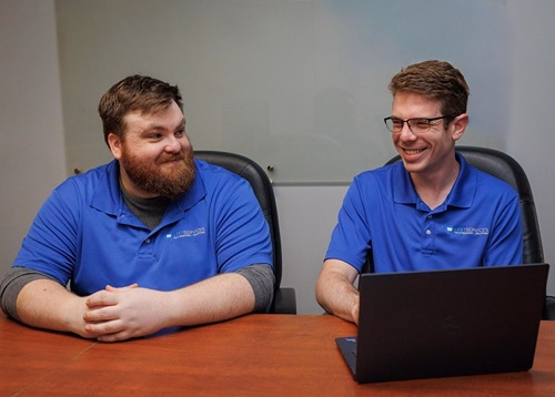 Two Leet IT support technicians in blue polo shirts smiling and collaborating on a laptop in a conference room