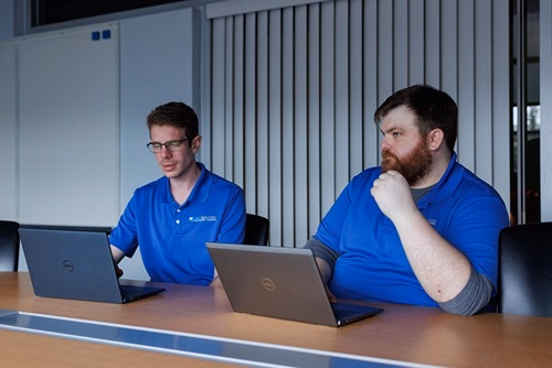 Two IT professionals in blue polo shirts working on Dell laptops at a conference table in a modern office