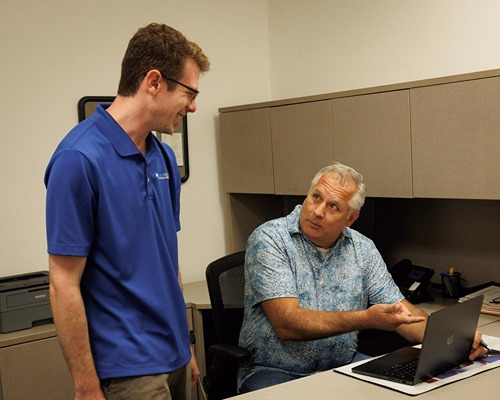 Two men discussing IT support solutions in an office, one seated at a laptop, one standing, having a professional conversa...