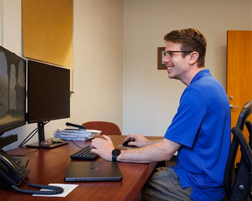 IT professional in blue polo shirt smiling while working at dual-monitor desktop workstation in office