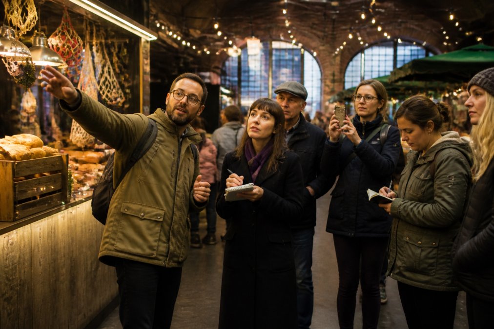 A knowledgeable food tour guide leading a small group of engaged participants through a vibrant Manchester food market, pointing out local producers and artisan food stalls, with participants taking notes and photos in natural market lighting