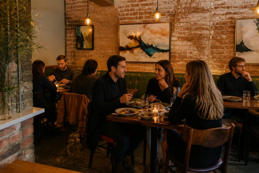 Intimate interior of a small independent restaurant with warm ambient lighting, wooden tables, locally sourced artwork on exposed brick walls, and diners enjoying small plates and conversation