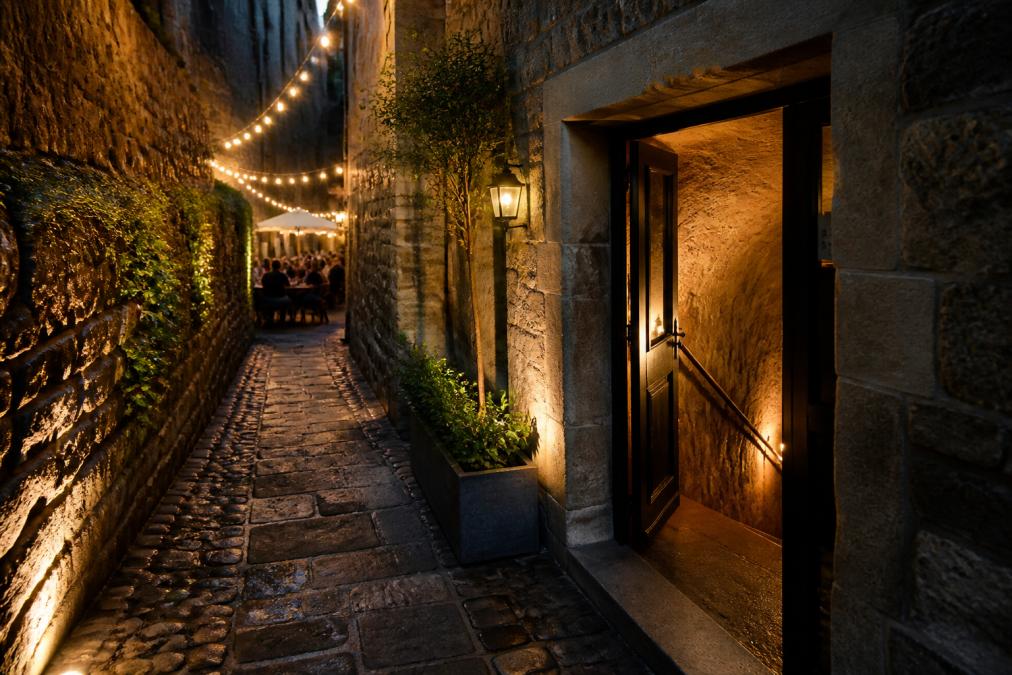 Narrow cobbled alleyway entrance to a hidden basement restaurant with warm light spilling from the doorway, atmospheric and inviting street-level view, evening lighting