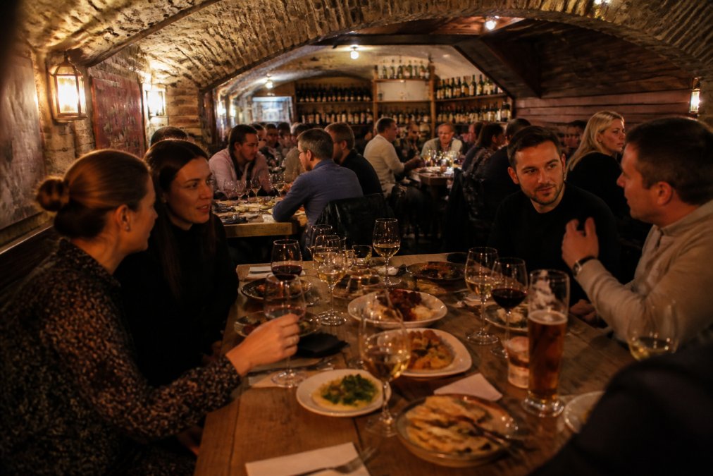 Intimate basement dining room with warm amber lighting, wooden tables crowded with small plates of Spanish tapas, glasses of wine and beer, and diners leaning in for conversation