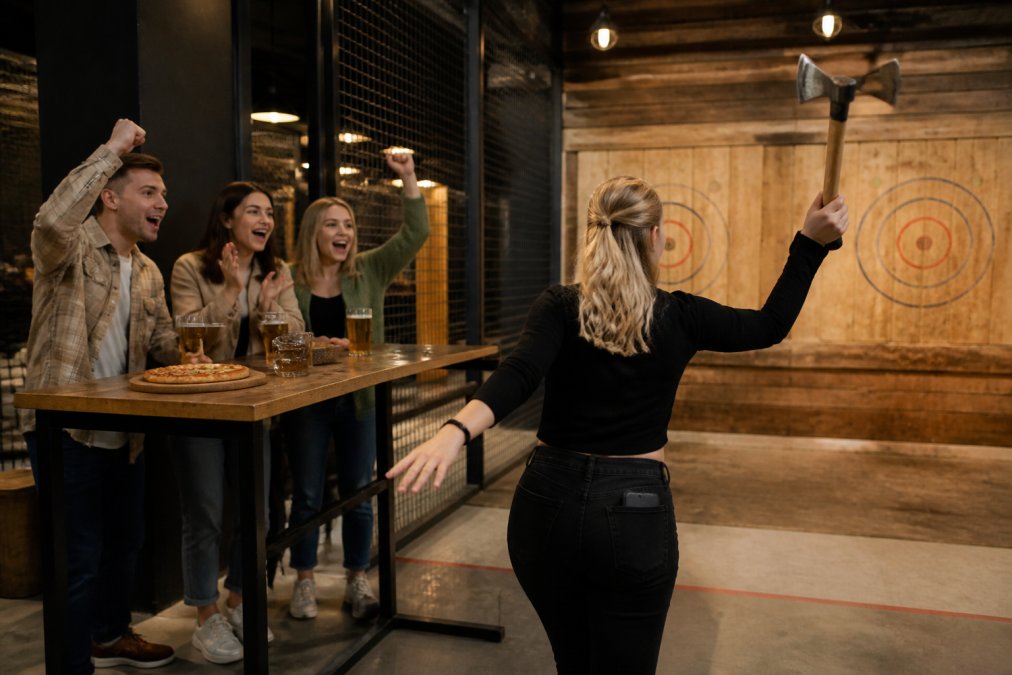 A participant in mid-throw at an axe throwing venue, concentrating intensely on a wooden target downrange, friends cheering enthusiastically at a nearby high-top table with craft beers and pizza, warm overhead lighting illuminating the throwing lanes