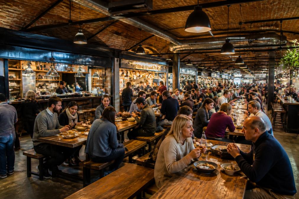 A vibrant indoor food hall with soaring brick ceilings and industrial architecture, independent food stalls visible in the background, diners seated at communal wooden tables sampling various cuisines, warm pendant lighting overhead