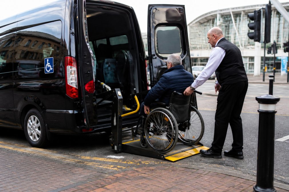 A wheelchair user boarding an accessible minibus or taxi at a designated drop-off point, with clear blue badge signage visible and the vehicle's hydraulic lift platform lowered, showing the accessible features and professional driver assistance