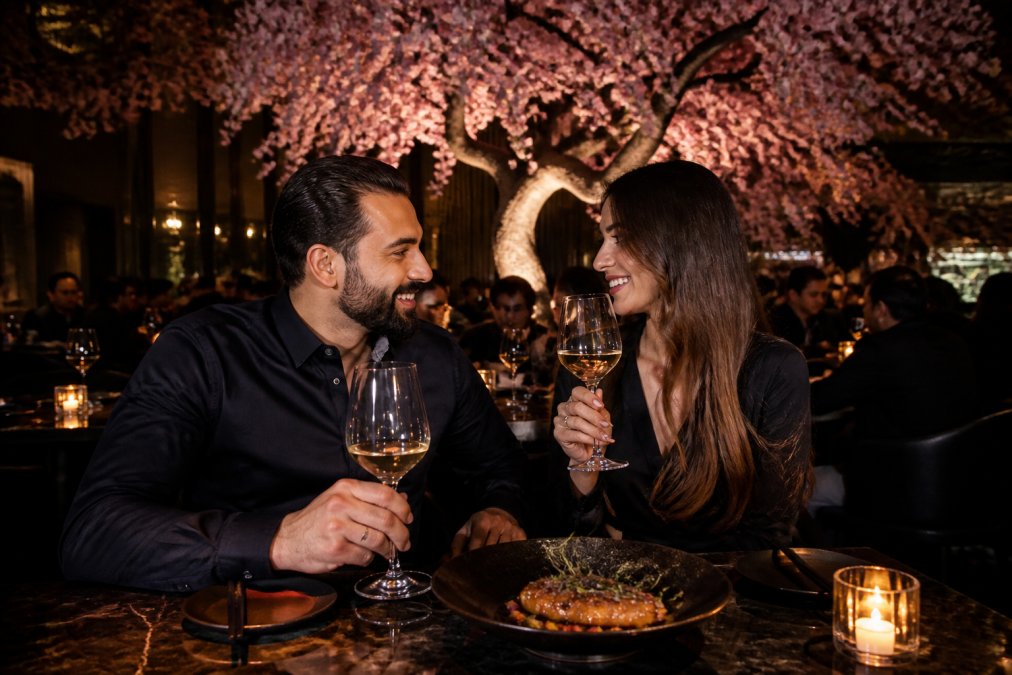 A couple seated at an intimate candlelit table in Tattu's dining room, the dramatic 40-foot cherry blossom tree installation glowing softly behind them, an elegantly plated modern Chinese dish in front, soft warm lighting reflecting off wine glasses