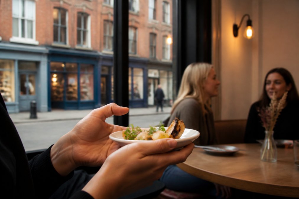 Close-up of hands holding a small plate of food at a tasting stop, with warm interior lighting of independent eatery visible in background, neighbourhood street scene through window showing brick buildings and local shopfronts