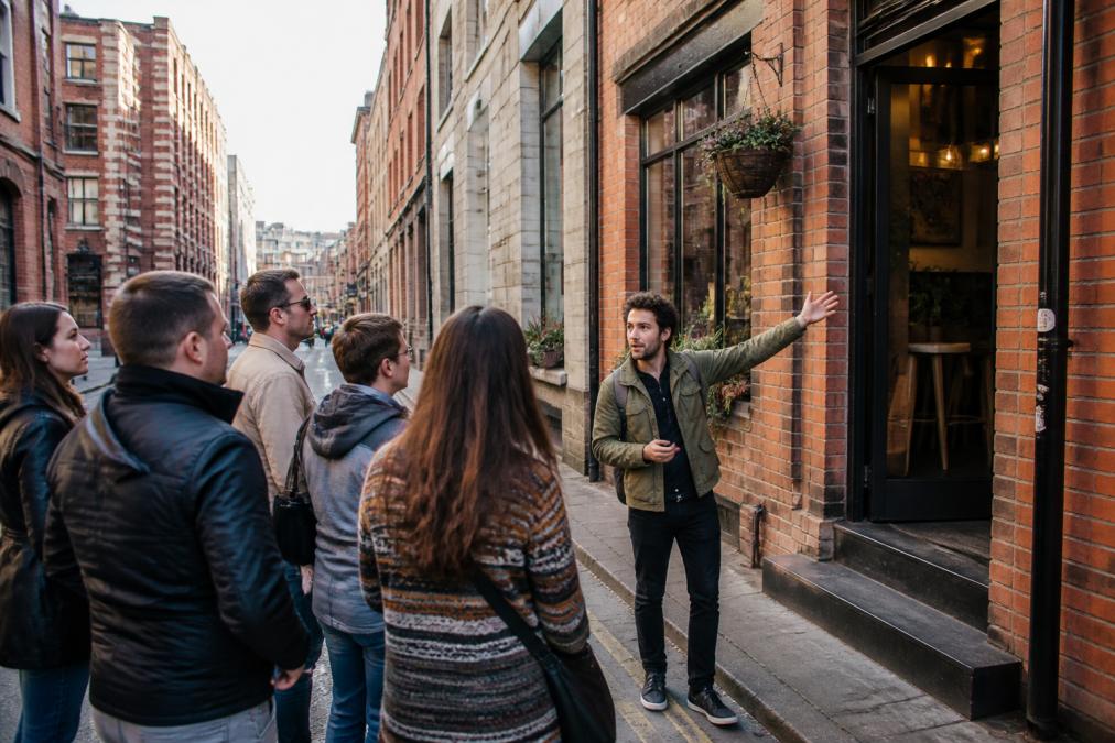 Small group of people walking together down a narrow urban street in Manchester's Northern Quarter, stopping outside an independent café with brick facade, a local guide gesturing toward the storefront entrance, afternoon light casting shadows on the pavement