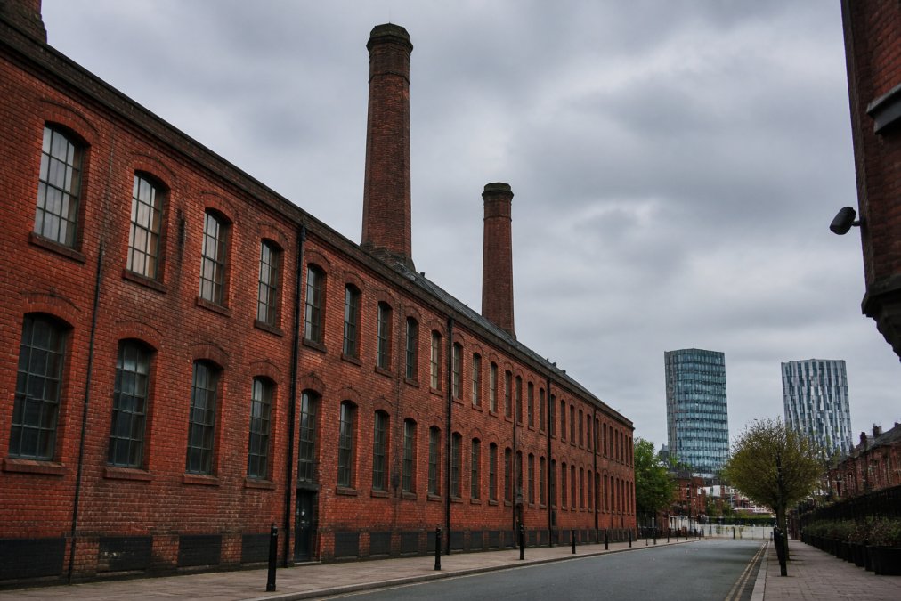 Historic brick cotton mill building with tall Victorian chimneys rising above surrounding roofline, weathered red brick facade with regular rows of tall windows, viewed from street level with modern city buildings visible in the distance
