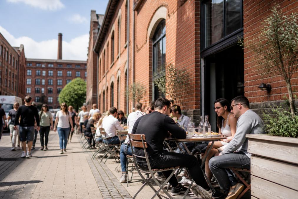 A lively street scene in Ancoats with converted red-brick mills and people dining outside an independent cafe on a sunny afternoon