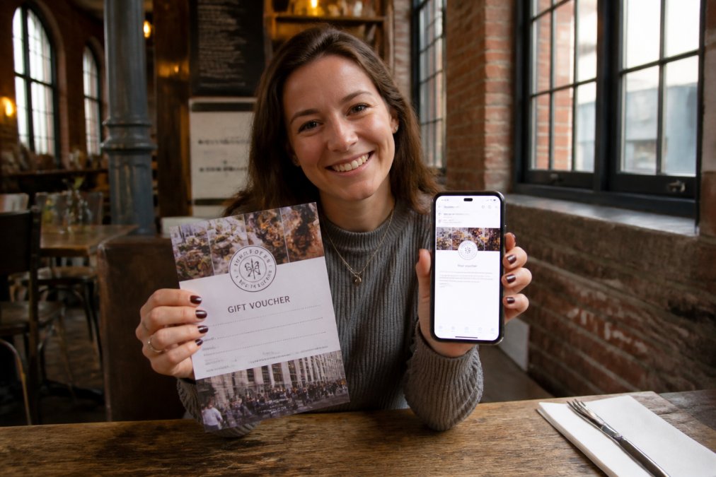 A person smiling as they hold a printed gift voucher beside a smartphone showing a digital e-voucher confirmation, seated at a cafe table in a historic Manchester building with exposed brick and large windows