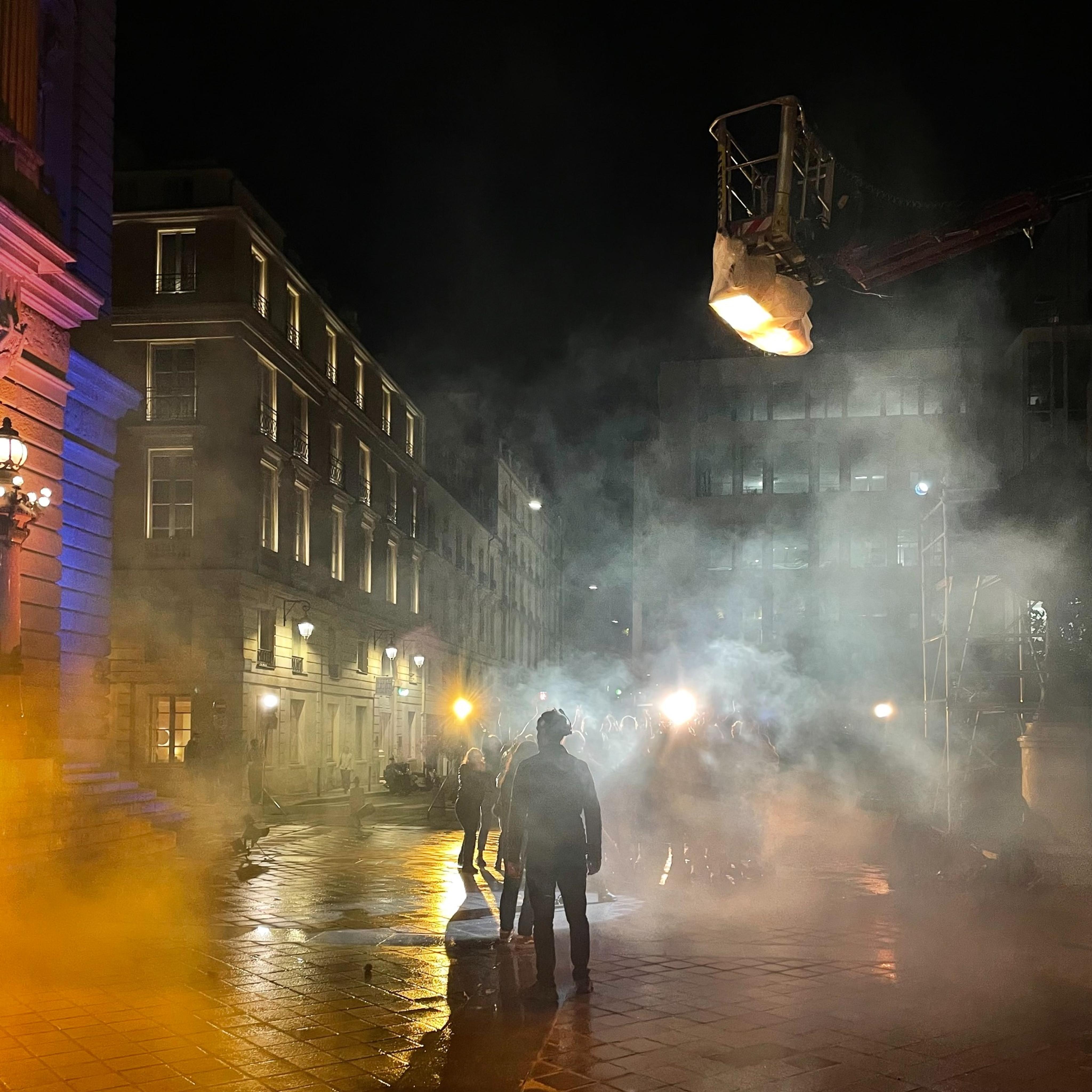 Nighttime film shoot on a misty French street, with crew silhouetted against bright lights and atmospheric smoke, showcasing Fixer France managing a cinematic urban set.
