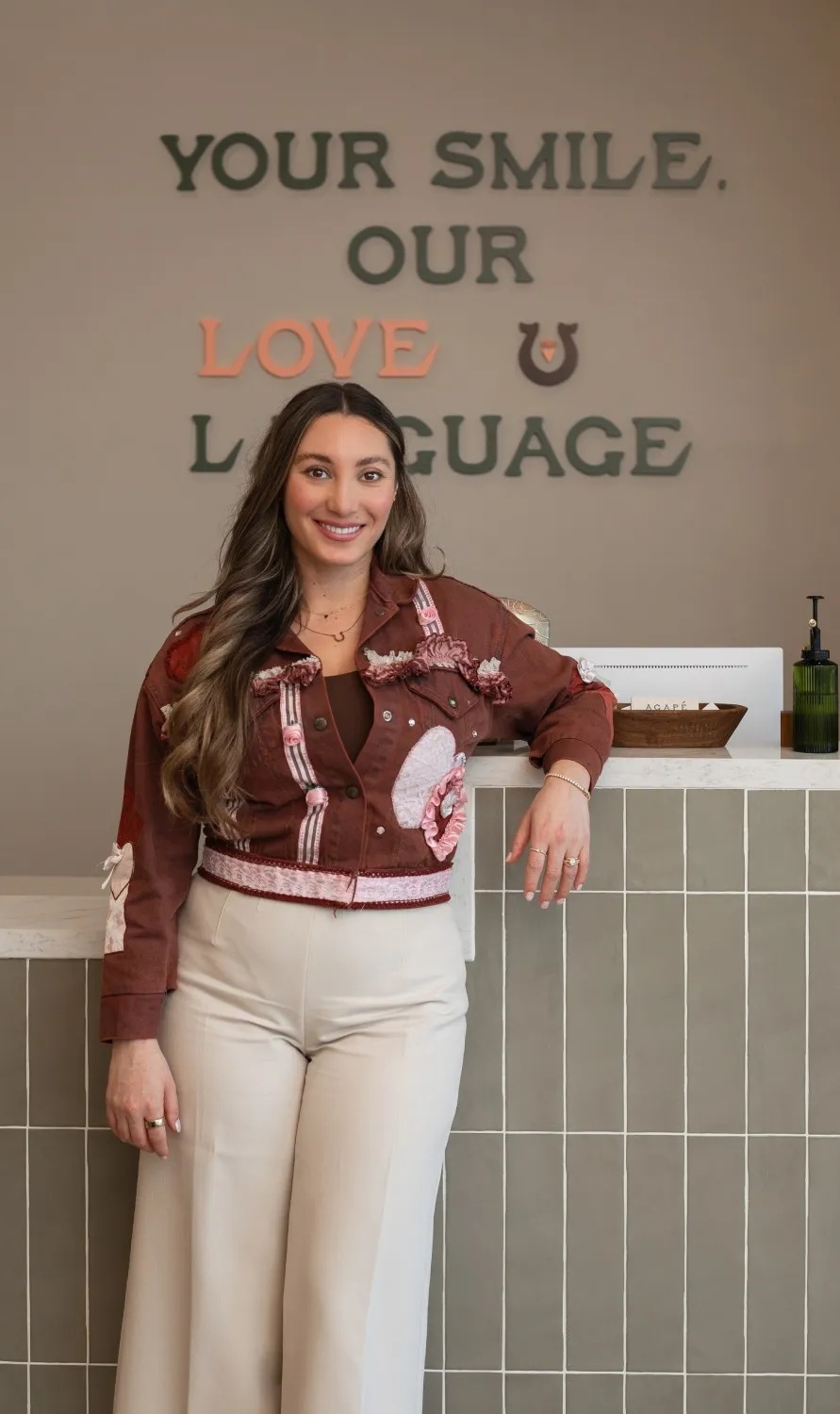 Smiling woman with long hair wearing a decorated brown jacket and beige pants leaning against a tiled counter under the text 'YOUR SMILE, OUR LOVE LANGUAGE'.