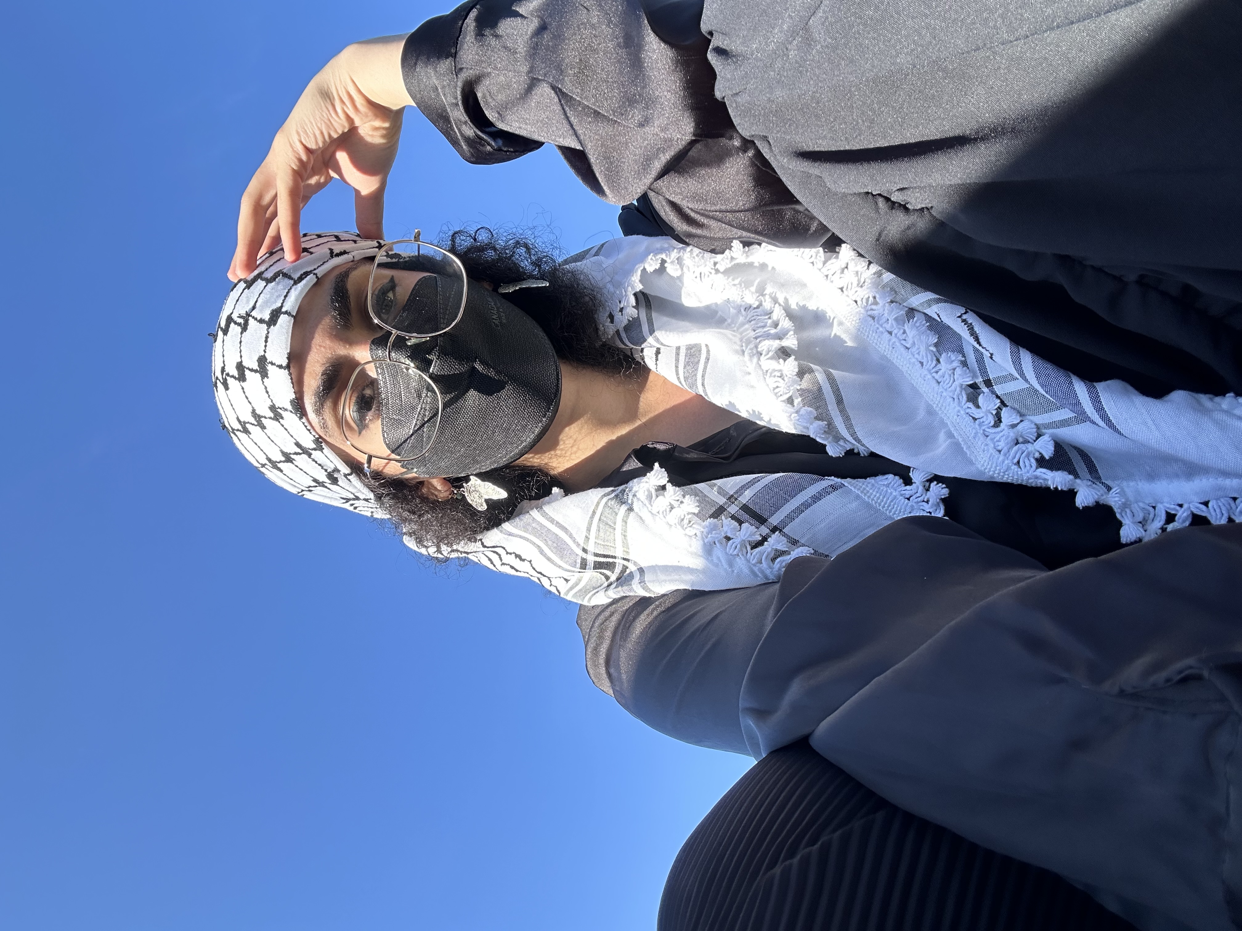 sama, an olive-skinned person wearing a keffiyeh, looks down at the camera. 