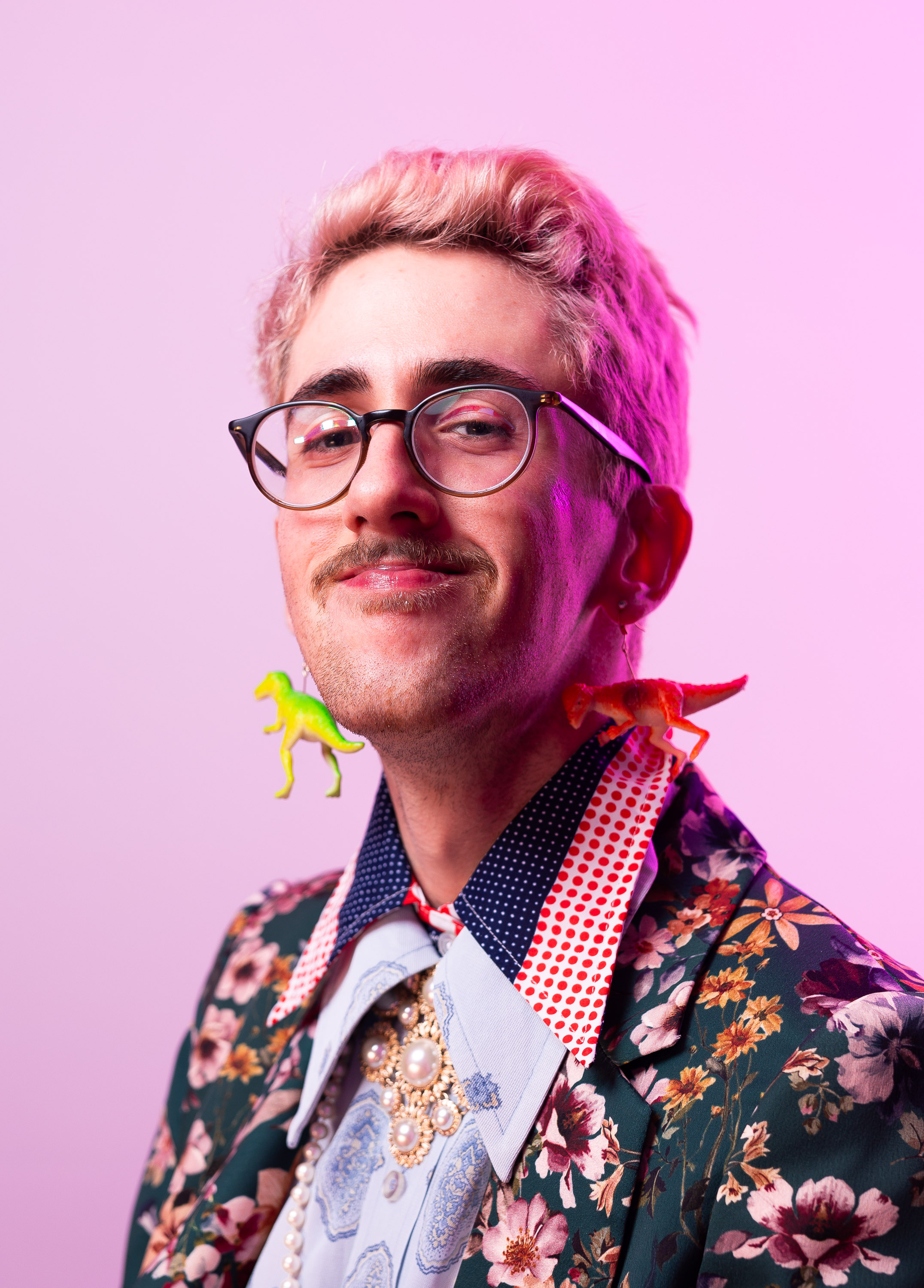 Liam smiles at the camera, with pink hair, facial hair, and round eyeglasses. 