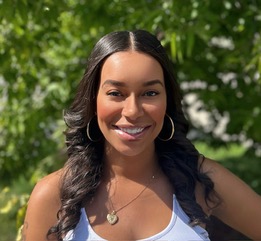 A Black woman stands in the sun and smiles big at the camera. 