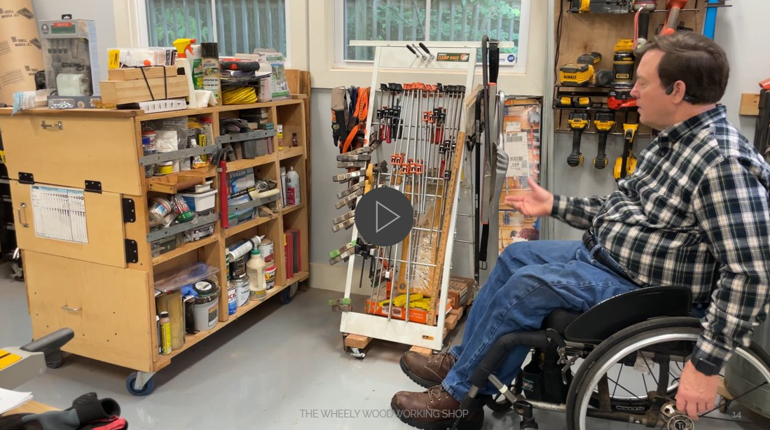 Man wearing black and white plaid shirt and jeans in manual wheelchair in front of his clamp rack and hardware racks, both mounted on casters so he can move them.