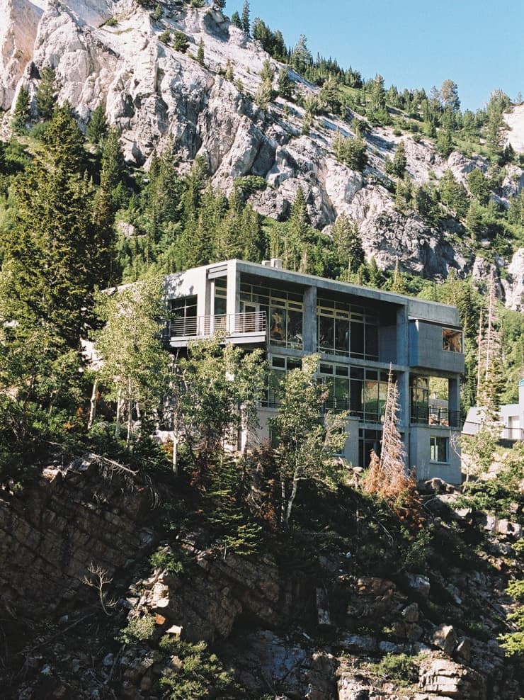 Modern concrete house with large windows nestled on a rocky hillside surrounded by evergreen trees and steep mountain cliffs.