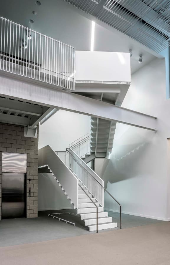 Modern indoor staircase with white railings and steps adjacent to a metallic elevator door in a minimalist building interior.