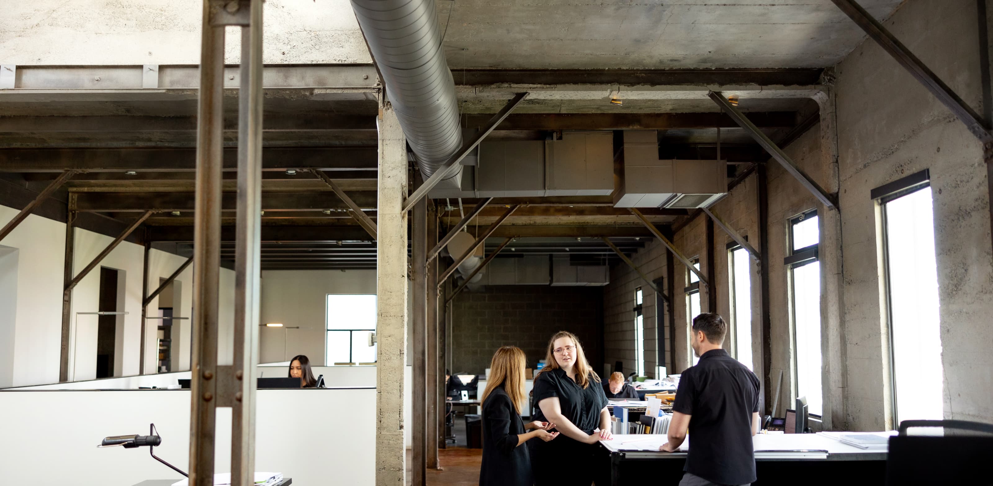 Three colleagues discussing over a large desk in an industrial-style office with concrete walls and exposed ductwork.