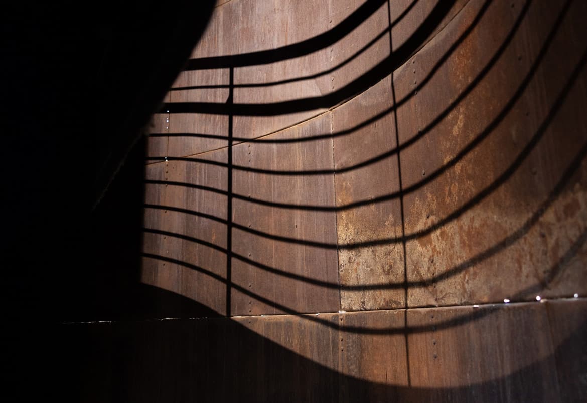 Curved metal wall with shadows of a railing creating a striped pattern across the surface.