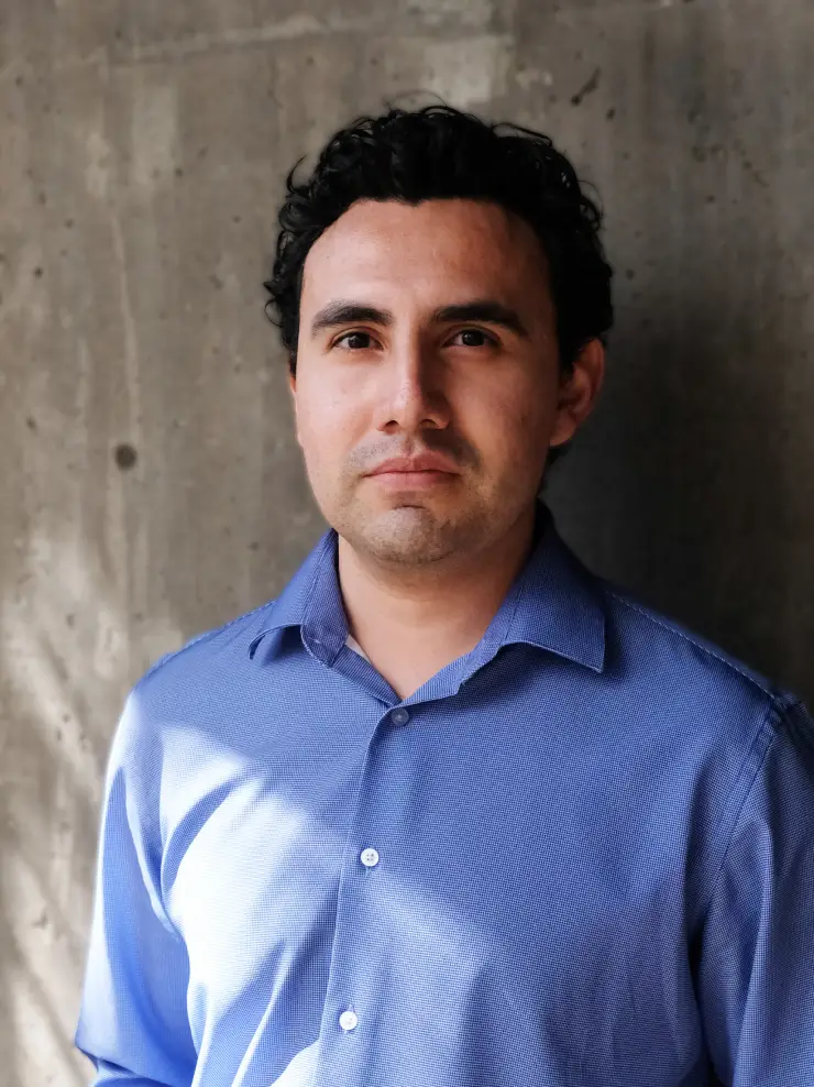 Man with short curly black hair wearing a blue button-up shirt standing against a concrete wall.