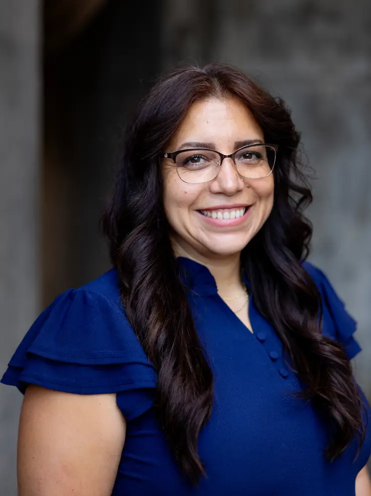 Smiling woman with long wavy dark hair, wearing glasses and a blue blouse with ruffled sleeves.
