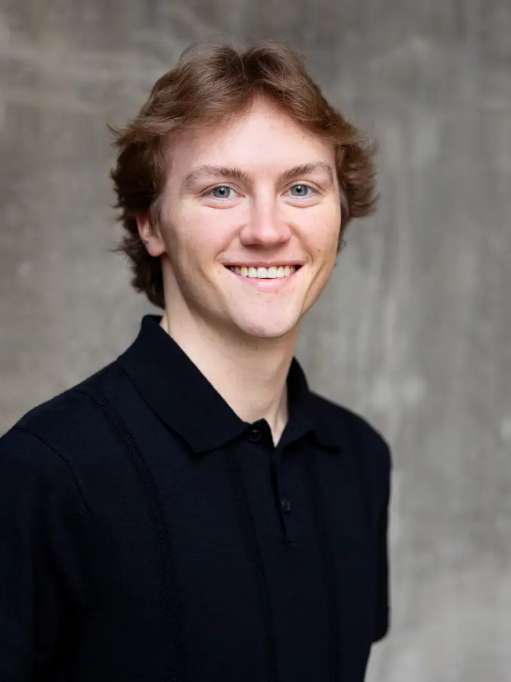 Young man with light brown hair and blue eyes smiling, wearing a black collared shirt, against a blurred gray background.