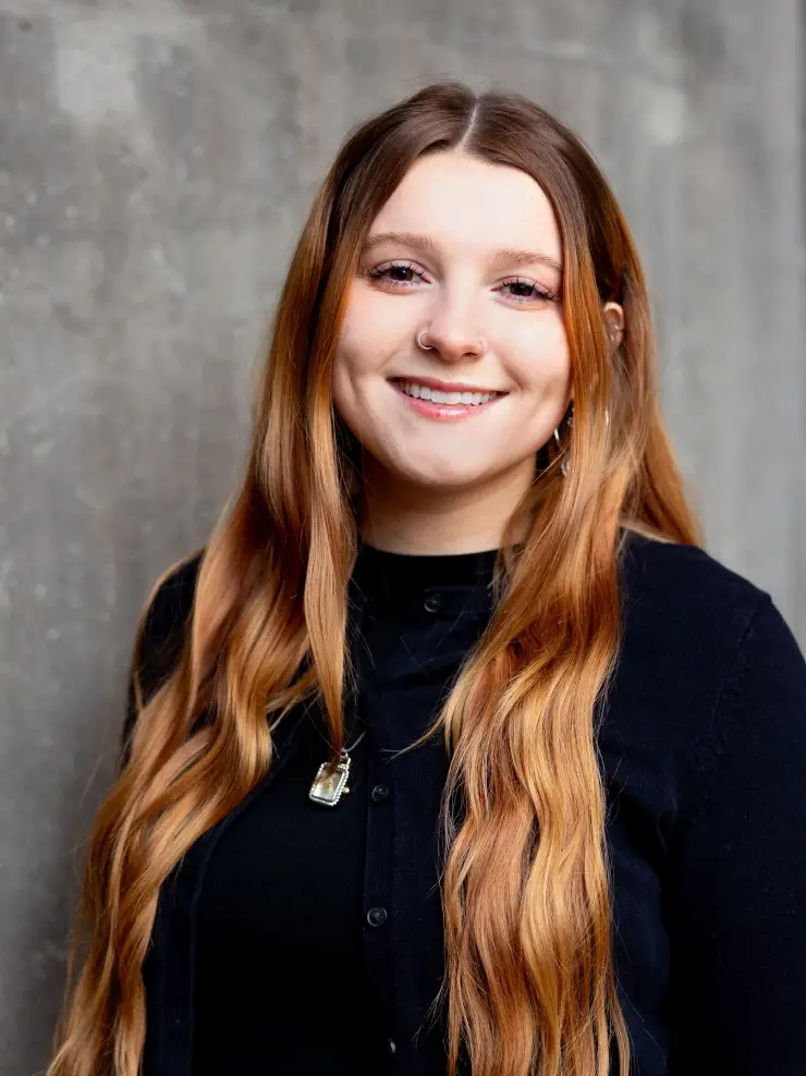 Smiling young woman with long wavy red hair wearing a black top and necklace against a gray background.