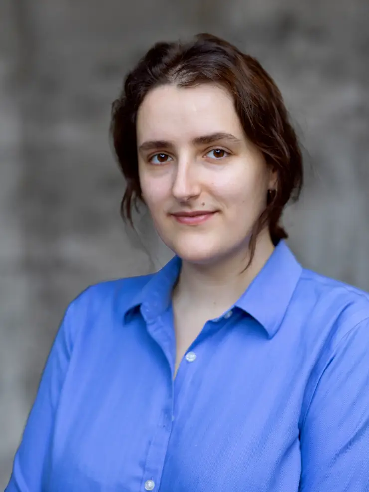 Portrait of a woman with brown hair wearing a blue button-up shirt against a gray blurred background.