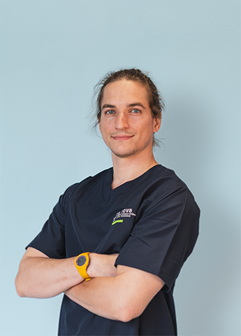 Man with long hair wearing a navy blue medical scrub top and yellow wristwatch, standing with arms crossed against a light blue background.